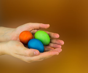 Three eggs lie in the palms of hands red green blue on a brown blurred background