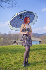 A young woman who is dressed to the latest fashion is holding a summer umbrella and is standing on the grass in a park.