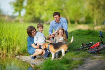 A full family is resting on the shore of a quiet lake. Mom, Dad, son, daughter and their dog sat by the water