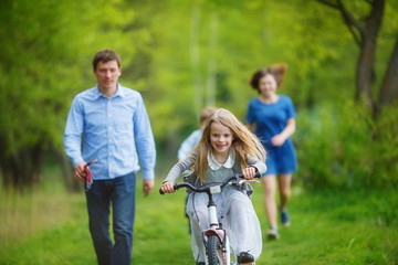 Family on a walk in the forest. A girl rushes on a bicycle on the green grass, parents try to catch up with her on foot