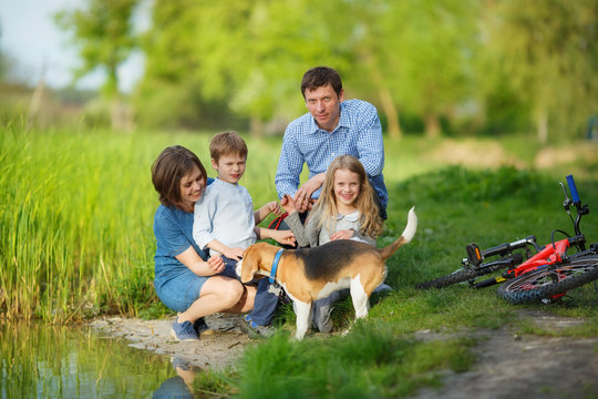 A Full Family Is Resting On The Shore Of A Quiet Lake. Mom, Dad, Son, Daughter And Their Dog Sat By The Water