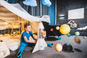 boy sitting in blanket fort with toy rocket and solar system model, father sitting on sofa behind © LIGHTFIELD STUDIOS