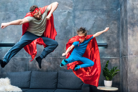 father and son in superhero costumes jumping on sofa at home