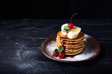 American pancakes on a plate with banana, cherry, mint caramel syrop and sugar powder. Dark background.