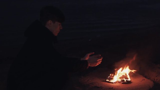 Young man seating near bonfire of burning shoes on the sand at coast strong bright flame summer dark night HD
