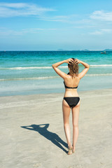 Back view of standing young blond woman. Young beautiful girl in black bikini relaxing on the tropical beach