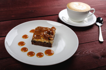 a piece of chocolate brownie and caramel sauce, a cup of cappuccino on a white plate. Top view
