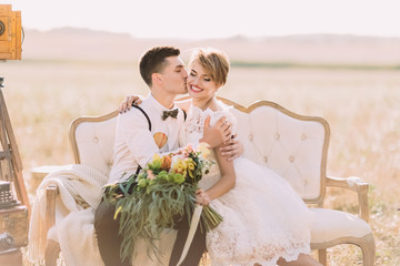 The romantic close-up portrait of the groom kissing the bride with the bouquet into the cheek in...