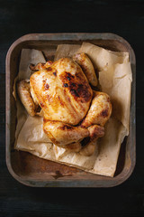 Grilled baked whole organic chicken on backing paper in old oven tray over black burnt wooden background. Top view, close up