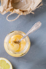 Jar of homemade lemon curd with spoon, whole and sliced lemons, sugar and zest over gray blue stone texture background. Top view with space.