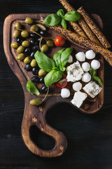 Mediterranean appetizer antipasti board with green black olives, feta cheese, mozzarella, capers, pepper, basil with grissini bread sticks over black wood burnt background. Top view with space © Natasha Breen