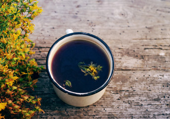 Herbal tea in an iron mug and St. John's Wort flowers on an old wooden surface of a table, soft focus