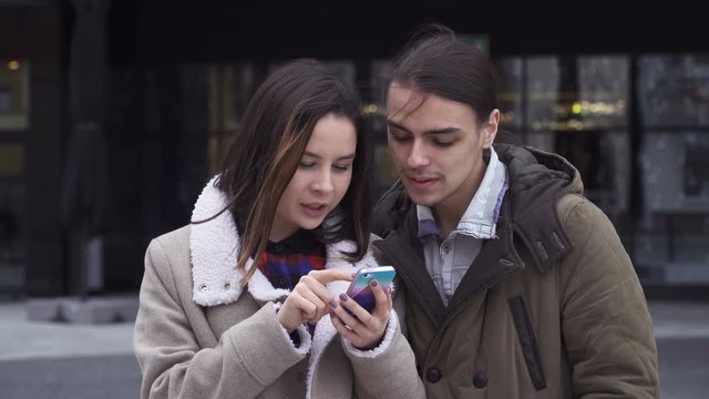 Young woman and man are looking at the screen of a cell phone, smiling and commenting emotionally what they see