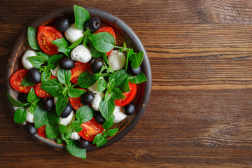 Classic Italian salad with mozzarella, tomatoes, basil, olives and olive oil. Caprese in a clay bowl on a wooden background. Top view, copy space.
