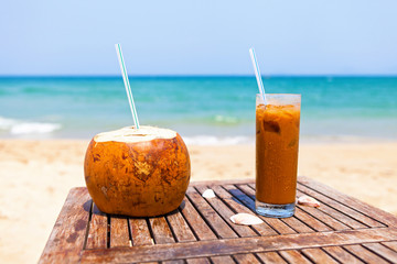 Coconut drink and ice coffee on the wooden table against beautiful beach and emerald sea