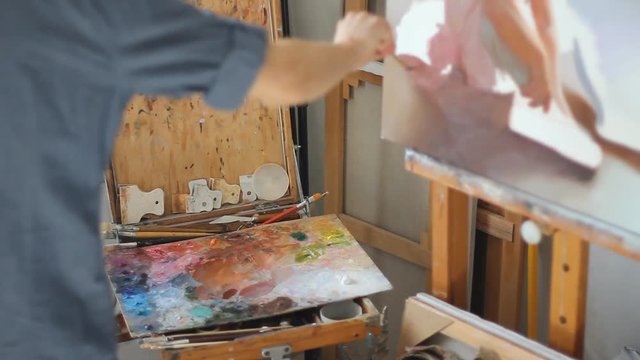 Studio workshop of a professional artist. Great set of pictures standing in a row near the wall. Graceful brush strokes. the man draws a little girl ballerina sitting on the floor. Baby clothes on the
