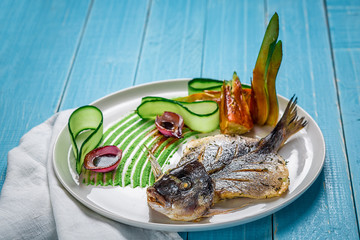 Fried fish dorado, decorated with cucumbers and onions. On a blue wooden background