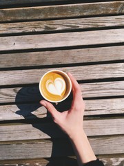 Woman hand holding a paper cup of coffee take away, heart sing from above. Latte cappuccino in cafe outside, wooden table