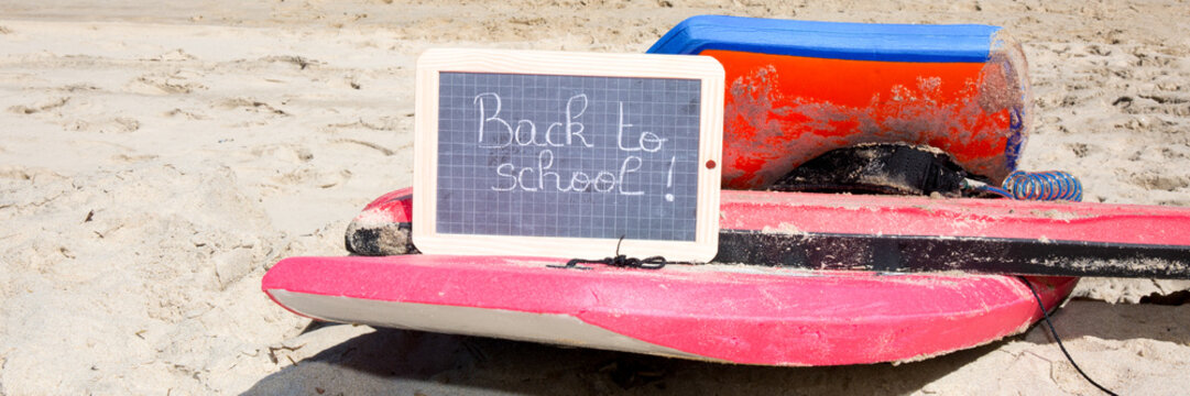 Surfing and bodyboarding are laid on the sand of the beach with a slate where is written back to school