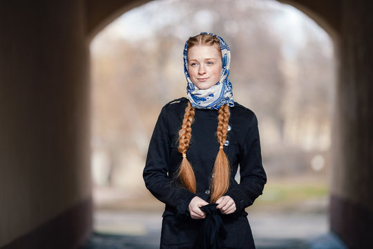 Young beautiful fashionable redhead woman with braids hairdo in blue white headcraft stylish denim black trench jacket posing outdoors in street arc spring time