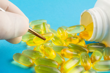 Hand in medical glove with tweezers keeps yellow capsule, lot of omega 3, fatty acid, fish oil on...