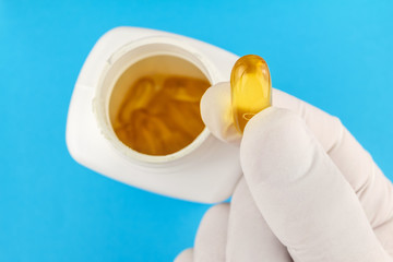 Hand in medical glove keeps yellow gel capsule of omega 3, fatty acid, fish oil, on blue background, white bottle, macro image.