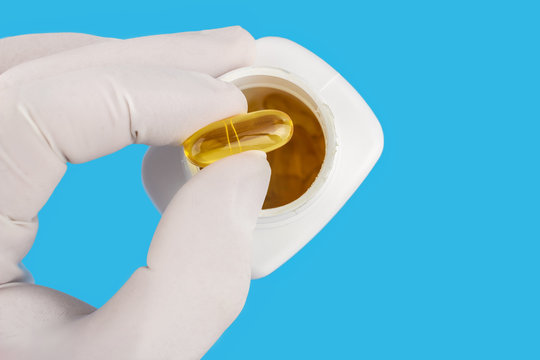 Hand in medical glove keeps yellow gel capsule of omega 3, fatty acid, fish oil, on blue background, white bottle, macro image.