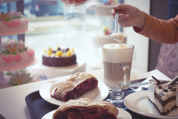 Child hand holding spoon above mug of latte among pieces of cherry strudel and cake.