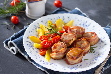 Rolls of meat in bacon and baked potatoes in rustic, fresh vegetables, tomato sauce and greens on a stone table.
