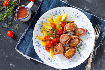 Rolls of meat in bacon and baked potatoes in rustic, fresh vegetables, tomato sauce and greens on a stone table.
