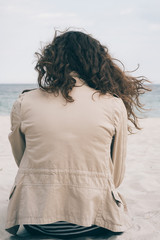 Girl with brown curly hair sits on the beach in windy weather