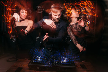 Two beautiful young girls dancing with handsome bearded disk jockey at DJ console. Party, dance,...
