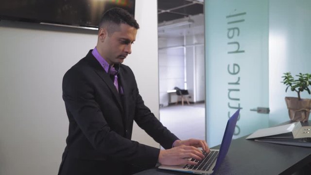 Portrait professional assistant working with laptop. Handsome man standing at reception desk entering data on computer. Focused worker looking on screen pc wearing in formal clothes