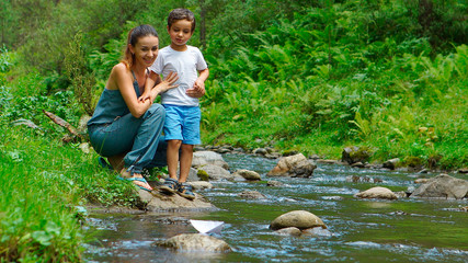 Mother with her son look at the toy paper boat on a stream
