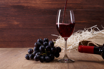 Red wine pouring. Wineglass with dark grapes branch. On rustic wooden background.