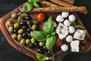 Mediterranean appetizer antipasti board with green black olives, feta cheese, mozzarella, capers, pepper, basil with grissini bread sticks over black wood burnt background. Top view with space