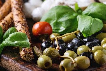 Mediterranean appetizer antipasti board with green black olives, feta cheese, mozzarella, capers, pepper, basil with grissini bread sticks over black wood burnt background. Close up