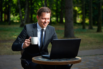 Handsome smiling businessman holding coffee cup in the park cafe next to his laptop.