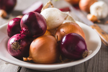 Several kinds of different onion bulbs at a white plate, standing on an old wooden table.