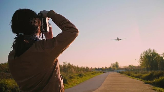 Rear view on young brunette photographer girl in leather pilot jacket with camera taking photos of big airplane landing in airport Spotting hobby enthusiast