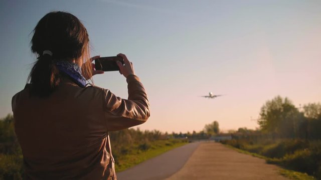 Rear view on young brunette photographer girl in leather pilot jacket with he smart phone taking photos and videos of big airplane landing in airport at morning sunrise time Spotting hobby enthusiast.