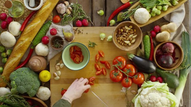 View from above of female hands cutting fresh tomatoes with knife on wooden cutting board and putting pieces into bowl. There is variety of fresh vegetables on kitchen table