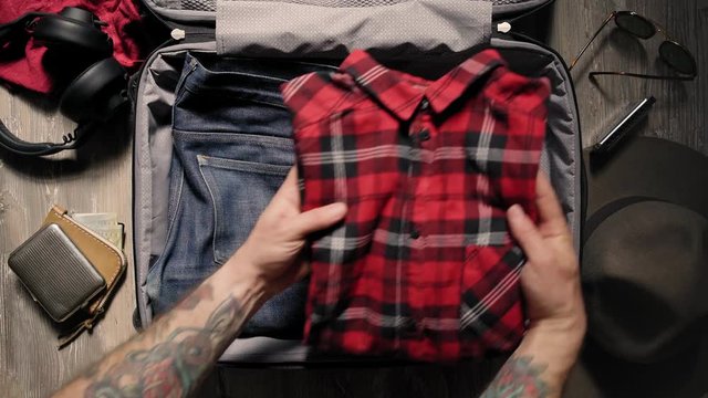 Manly young caucasian with strong arms with tattoos packs his suitcase full of manly artisanal items like selvedge denim, flannel shirt, stainless flask and other hipster accessories