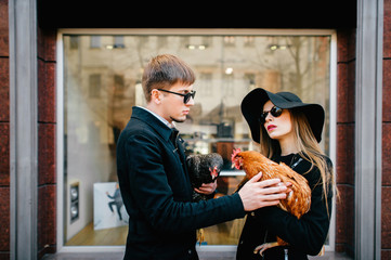 Portrait of beautiful couple of models in love in fashion clothes, black sunglasses, elegant hat standing in front of boutique showcase with chickens in hands and looking around. Unusual pets concept.