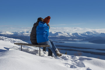 Fototapeta na wymiar Rear view of couple sitting on wooden bench, enjoying winter view of ocean and snow mountains in Norway.