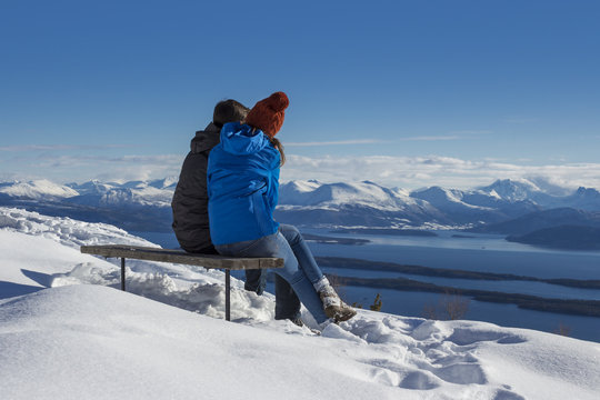 Rear view of couple sitting on wooden bench, enjoying winter view of ocean and snow mountains in Norway.