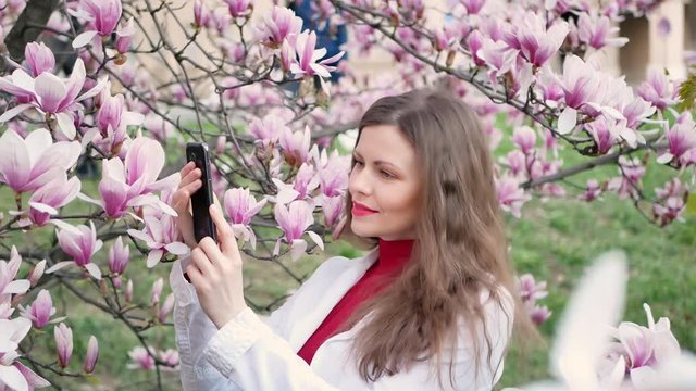 beautiful girl takes photos of magnolia pink blossom sping tree flowers on a smartphone