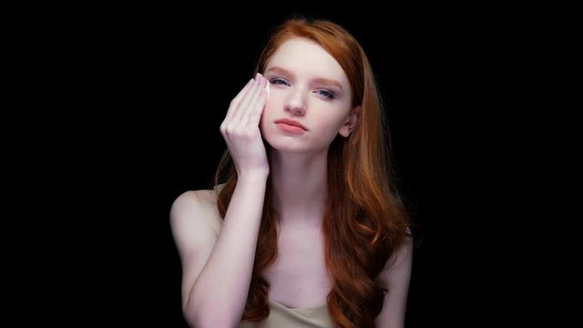 Beautiful young red haired woman cleaning her face with cotton pad isolated on black background
