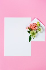 Colorful spring flowers in envelope and white sheet on pink background.