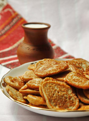 Heap of potato flapjacks, clay jug with milk and bowl with sour cream on the tablecloth with national pattern close up